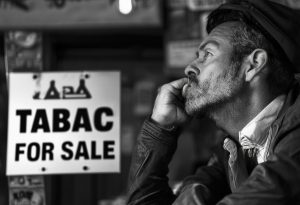 Image en noir et blanc montrant un entrepreneur ambitieux, pensif devant une enseigne 'Tabac à vendre', éclairage ambiant.