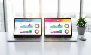 Un bureau moderne avec deux ordinateurs portables affichant des tableaux de bord colorés marqués KPI et OKR devant de grandes fenêtres baignées de lumière naturelle.